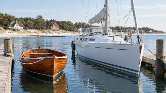 Kleines lackiertes Holzboot und eine weiße Segelyacht an einem Holzsteg, dahinter Strand, Kiefern und Häuser